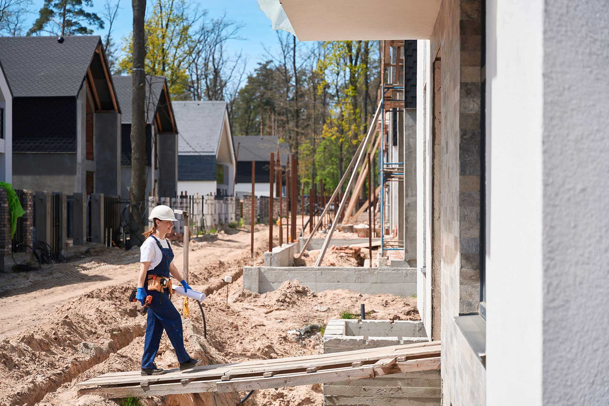 A construction worker, wearing a safety helmet and gear, walks on a wooden plank in a residential building site surrounded by unfinished houses.