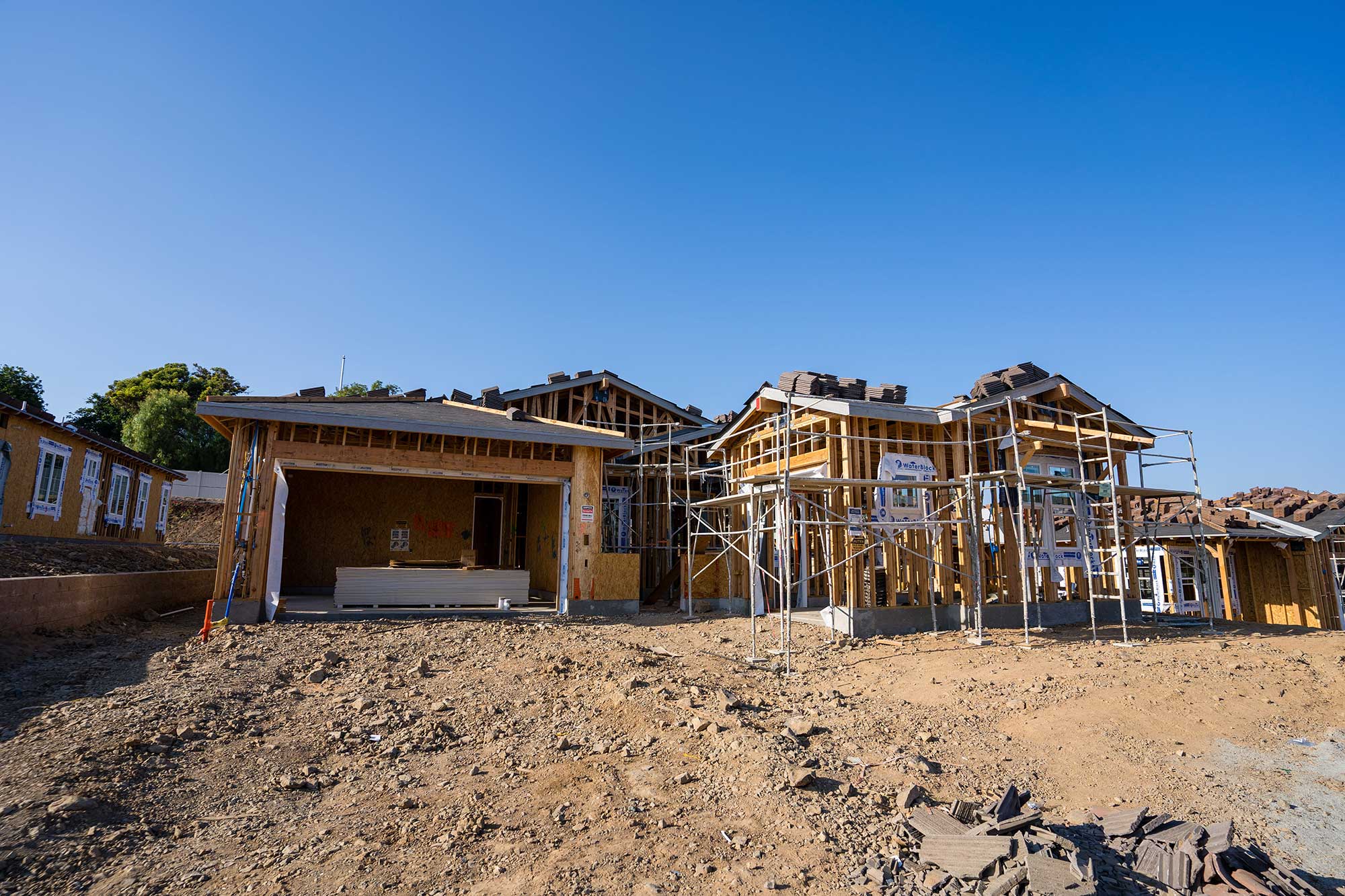 A partially constructed home with wooden framing, scaffolding, and roof tiles, set against a clear blue sky and rocky terrain.