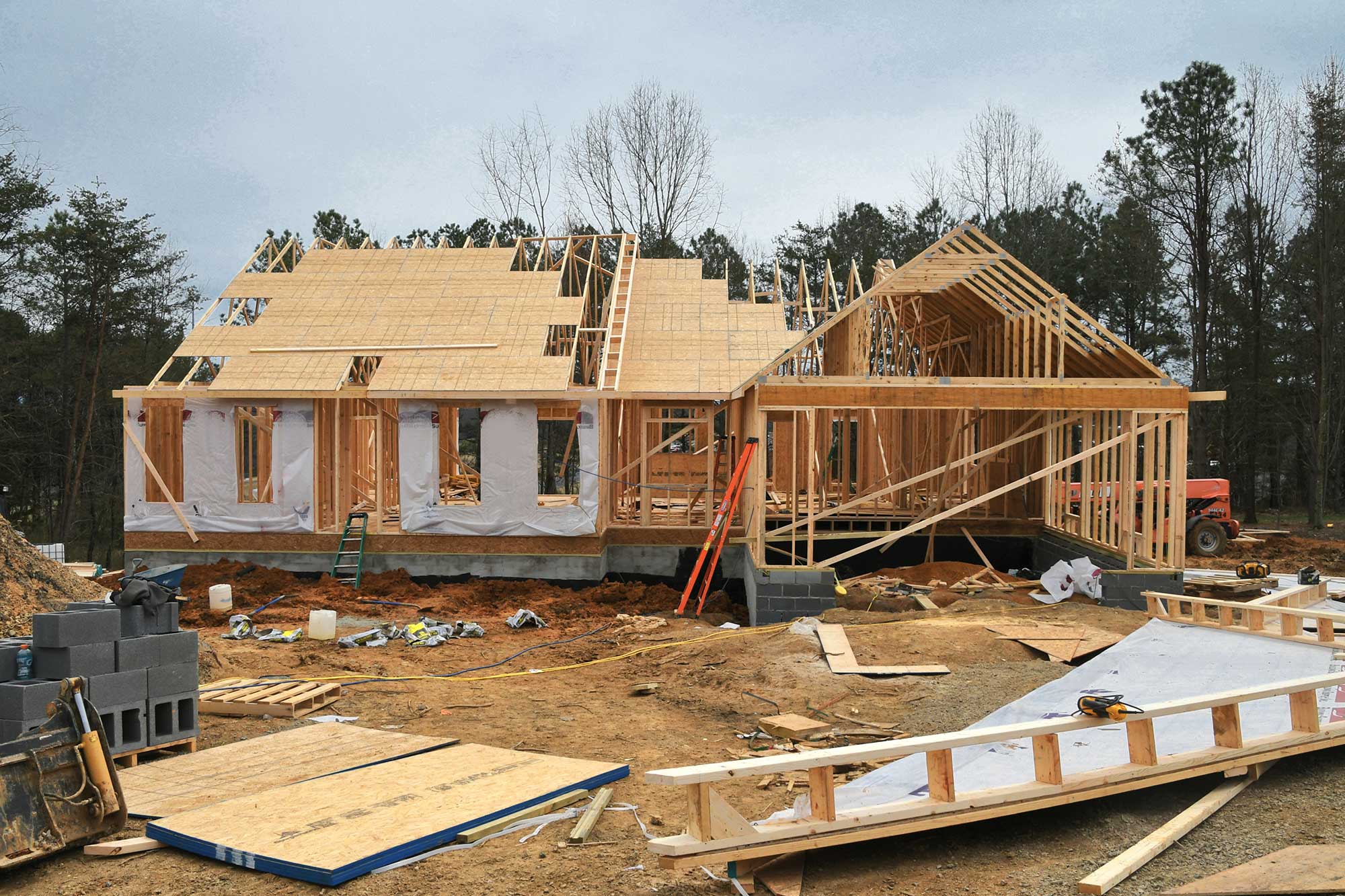 A partially constructed wooden house with exposed framing, stacks of building materials, and machinery on a cloudy day.