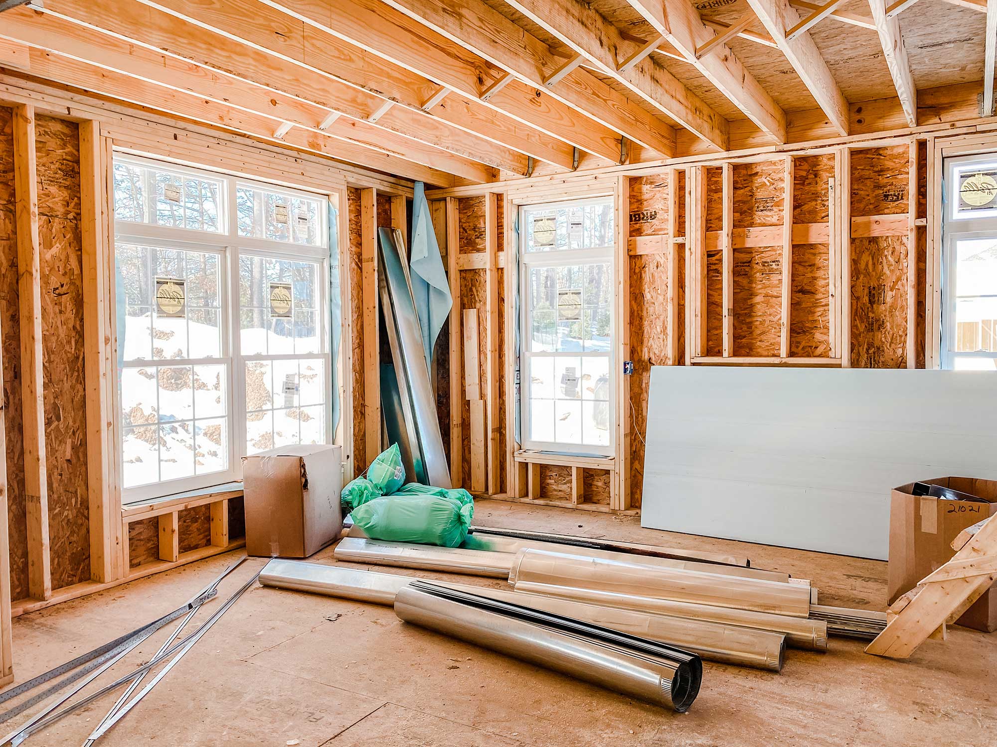 Bright, unfinished room with wooden framework, two windows, construction materials like sheets and pipes scattered on the floor.