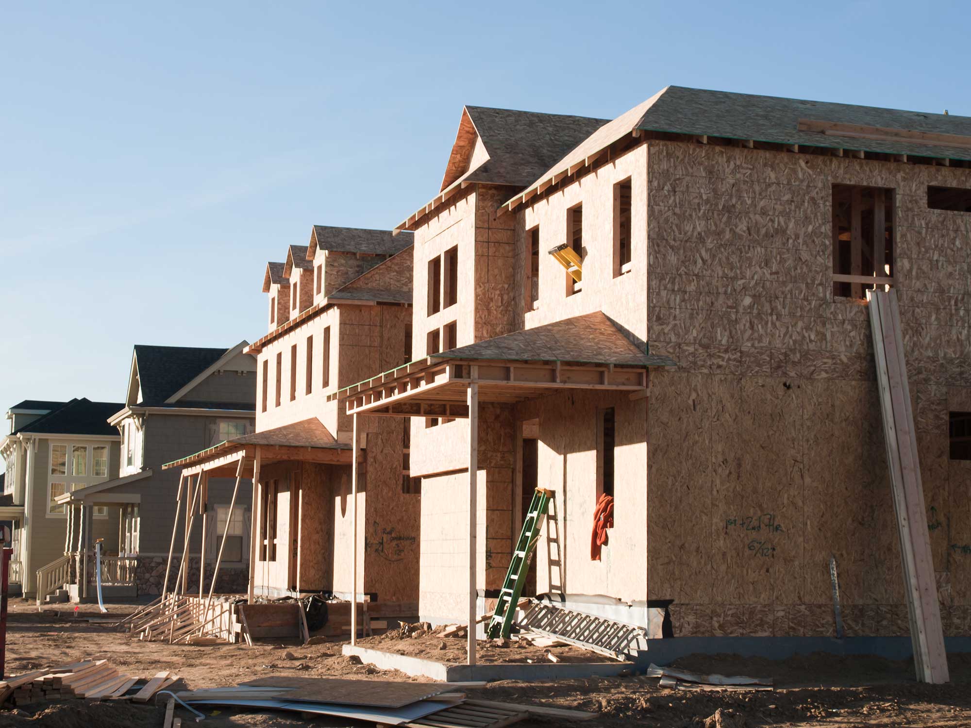 Construction site featuring partially built homes with wooden frames, stacks of lumber, and tools under a clear blue sky.