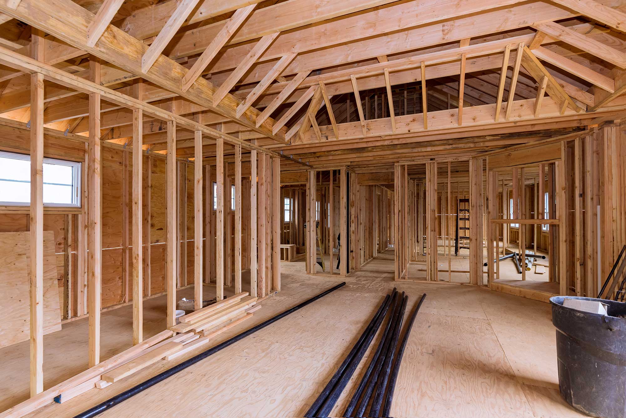 Interior of a framed house under construction, showcasing wooden beams, studs, and flooring with construction materials scattered around.