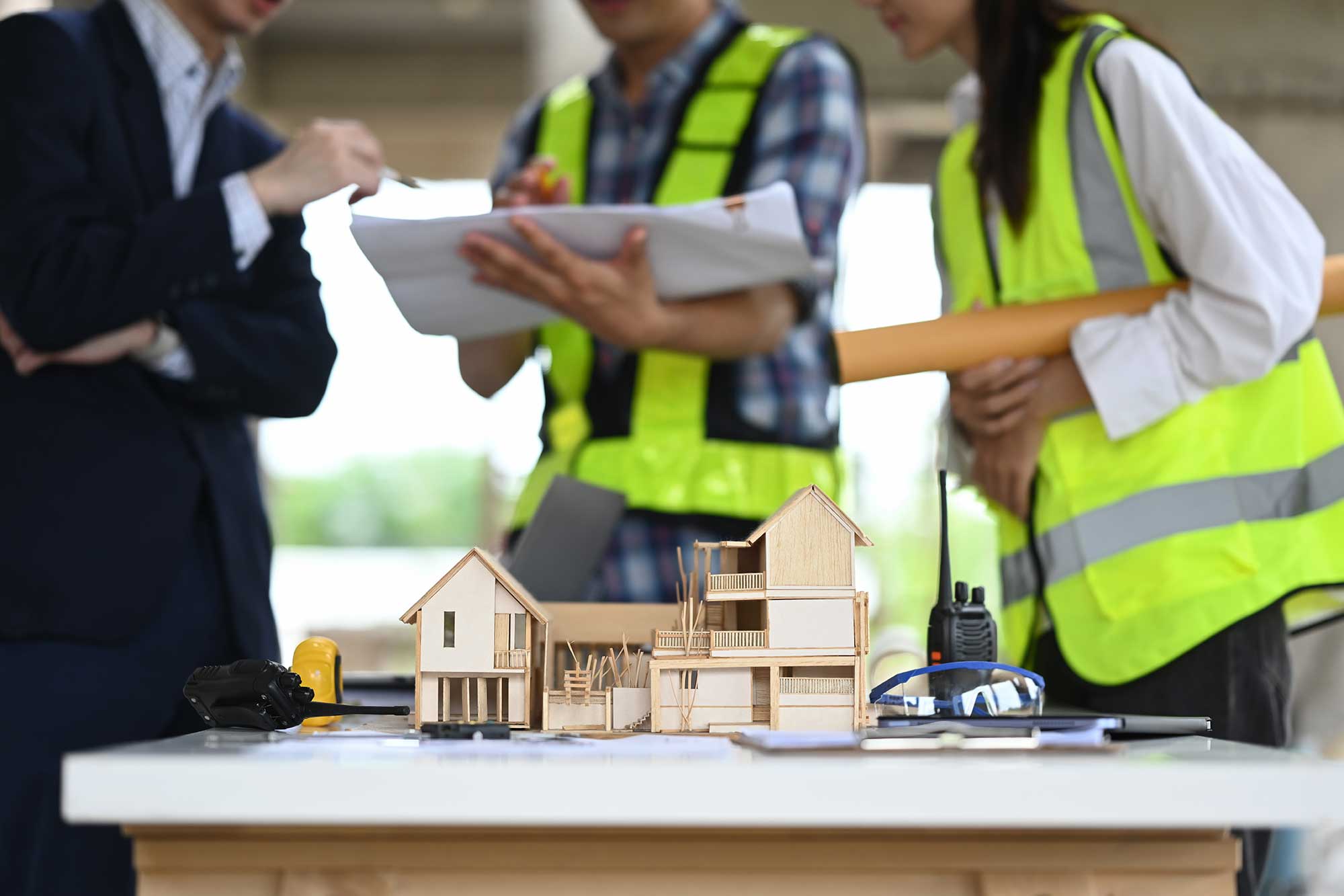Three construction professionals discuss plans over a wooden house model on a table, wearing safety vests, amidst a construction site.