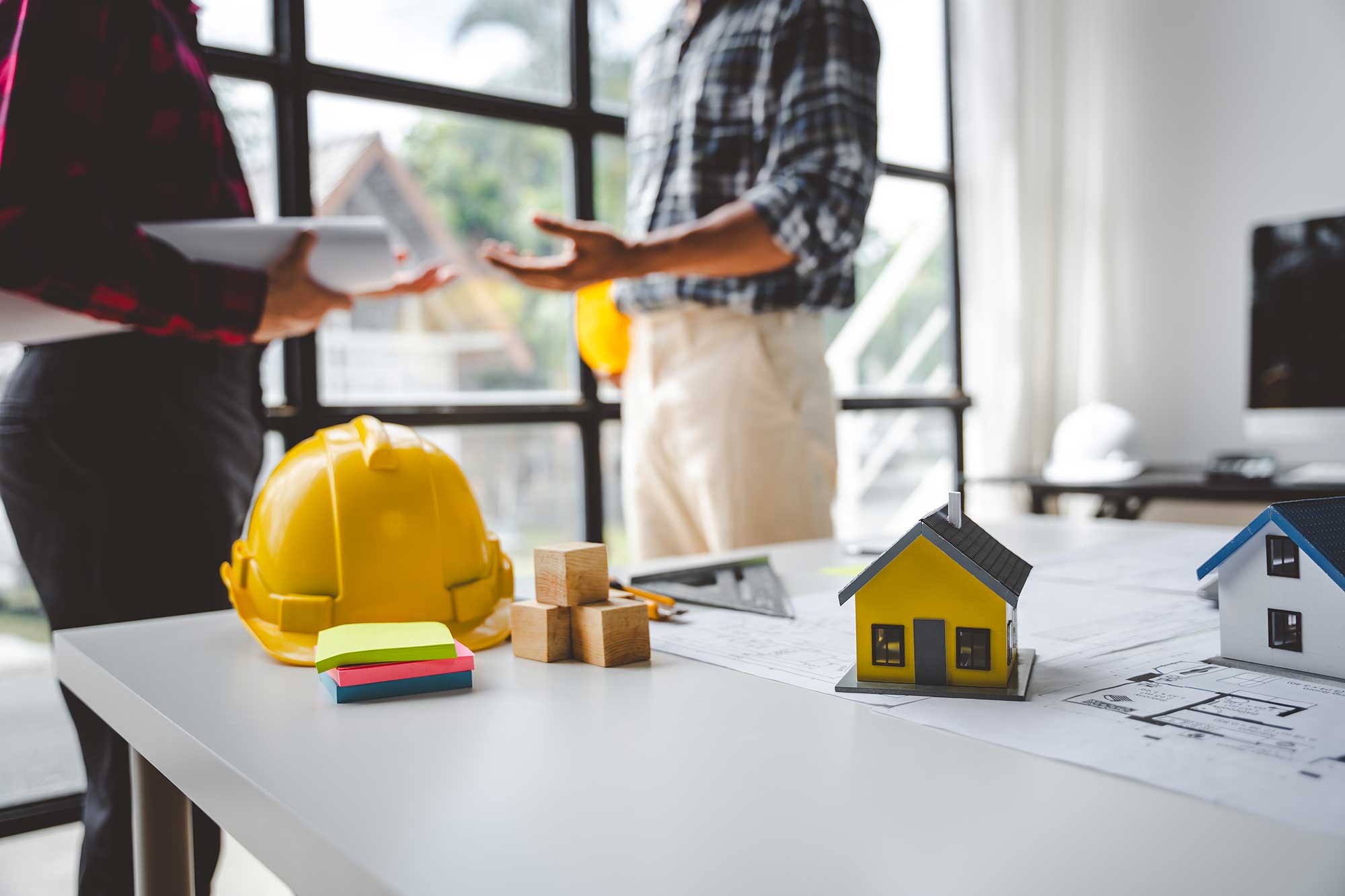 Two construction professionals discuss plans at a table, with yellow hard hats and miniature houses in a well-lit workspace.