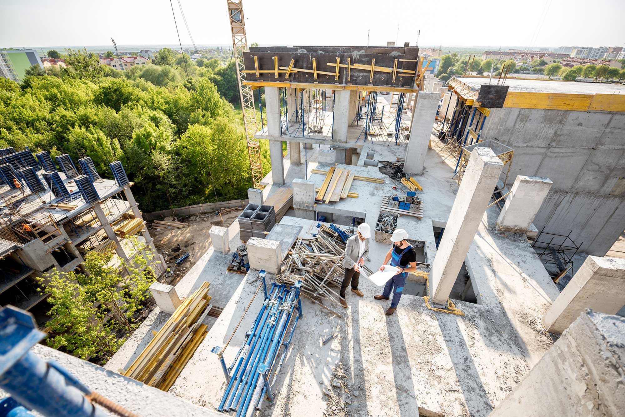 Two construction workers examine blueprints on a building site surrounded by greenery and urban development under clear skies.